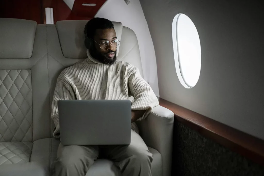 A man wearing glasses and a sweater sits comfortably in a plush leather seat inside a private jet, using a laptop and looking out of the window.